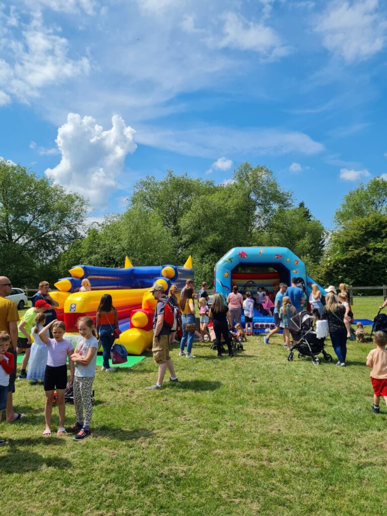 image shows lots of families with young children enjoying playing on a bouncy castle in the sunshine at a community event