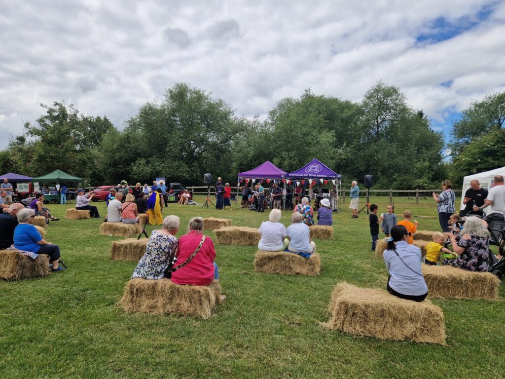 Image shows people sitting in a paddock at an event on hay bales watching a ukulele band perform